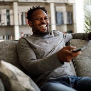 Man smiles while holding a remote, sitting on a gray couch in a living room with bookshelves and a plant.