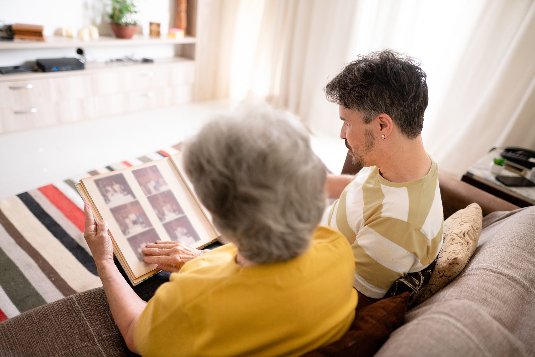Two people sit closely, looking at a photo album together in a cozy living room with a striped rug and soft lighting.