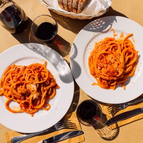 Two plates of pasta sit on a sunny table with red sauce, surrounded by a carafe of wine, glasses, cutlery, and a bread basket.