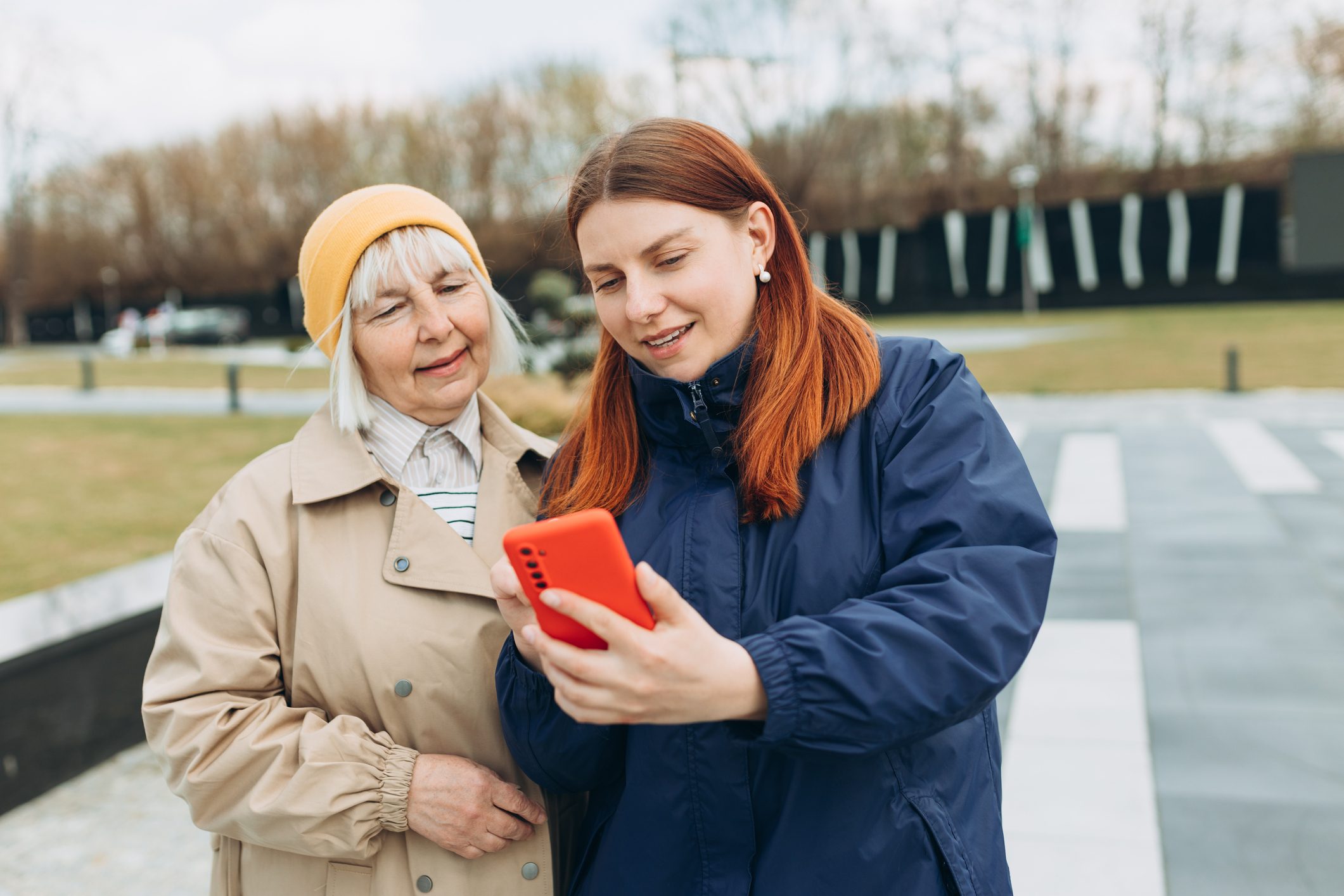 Two people look at a smartphone together in an outdoor setting, with trees and pathways in the background.