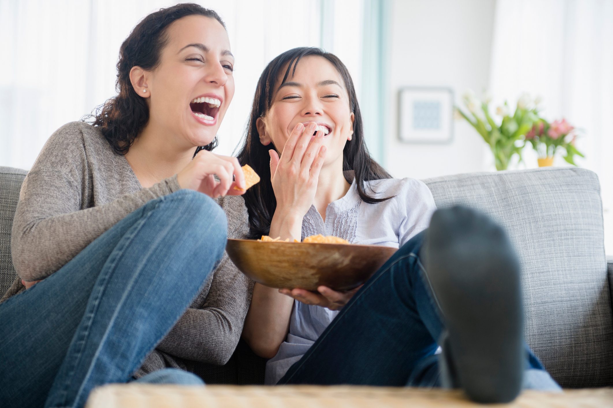 Two women laugh while eating chips from a bowl, sitting comfortably on a sofa in a bright, cozy living room.