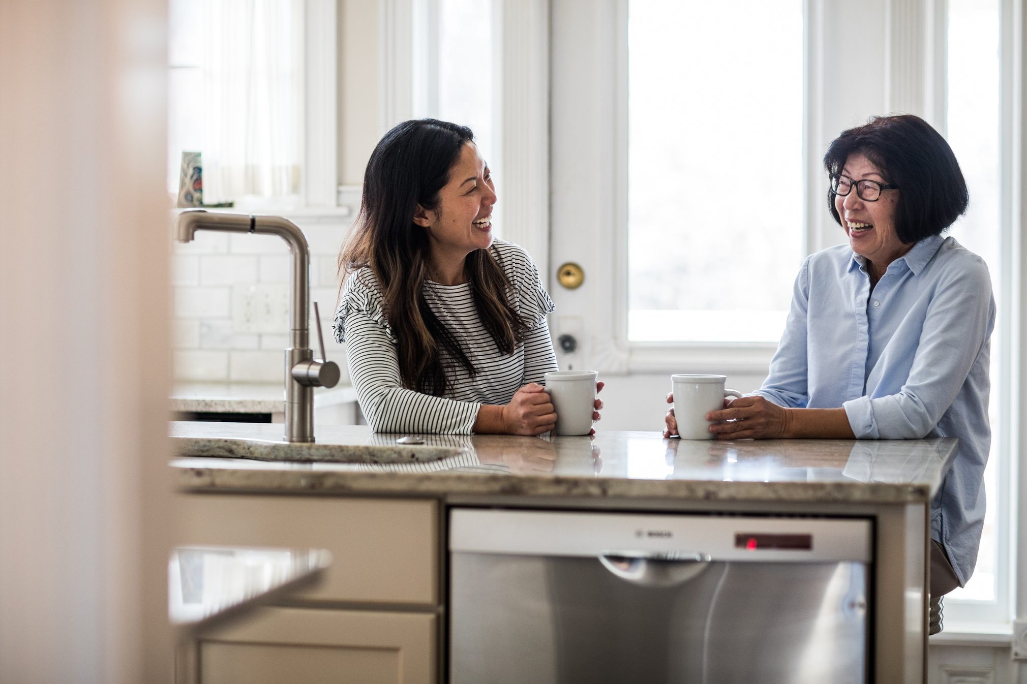 Two women sitting, laughing and drinking coffee at a kitchen counter, with a bright window in the background.