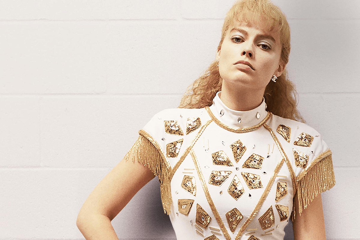 Person stands confidently, wearing a gold and white embellished costume, against a plain white wall, exuding determination and elegance.