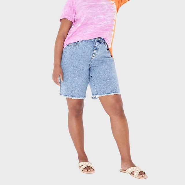 Person standing, wearing a pink and orange tie-dye shirt, denim shorts, and beige sandals; posing against a plain white background.
