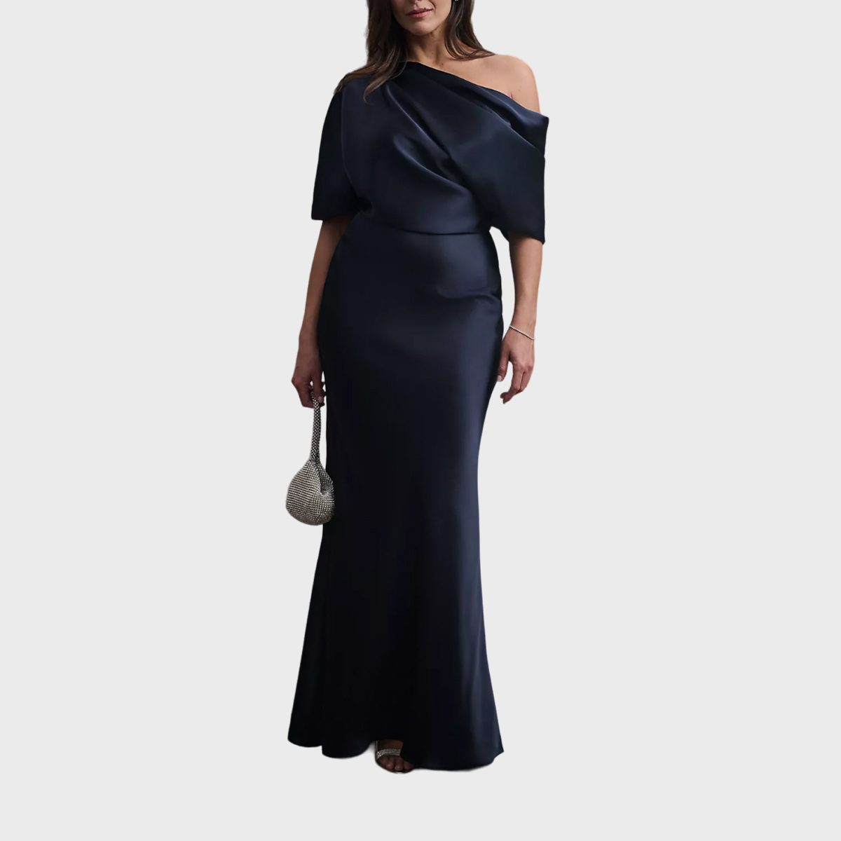 Woman wearing a dark, off-shoulder gown; holding a small, shimmering purse; standing against a plain background.
