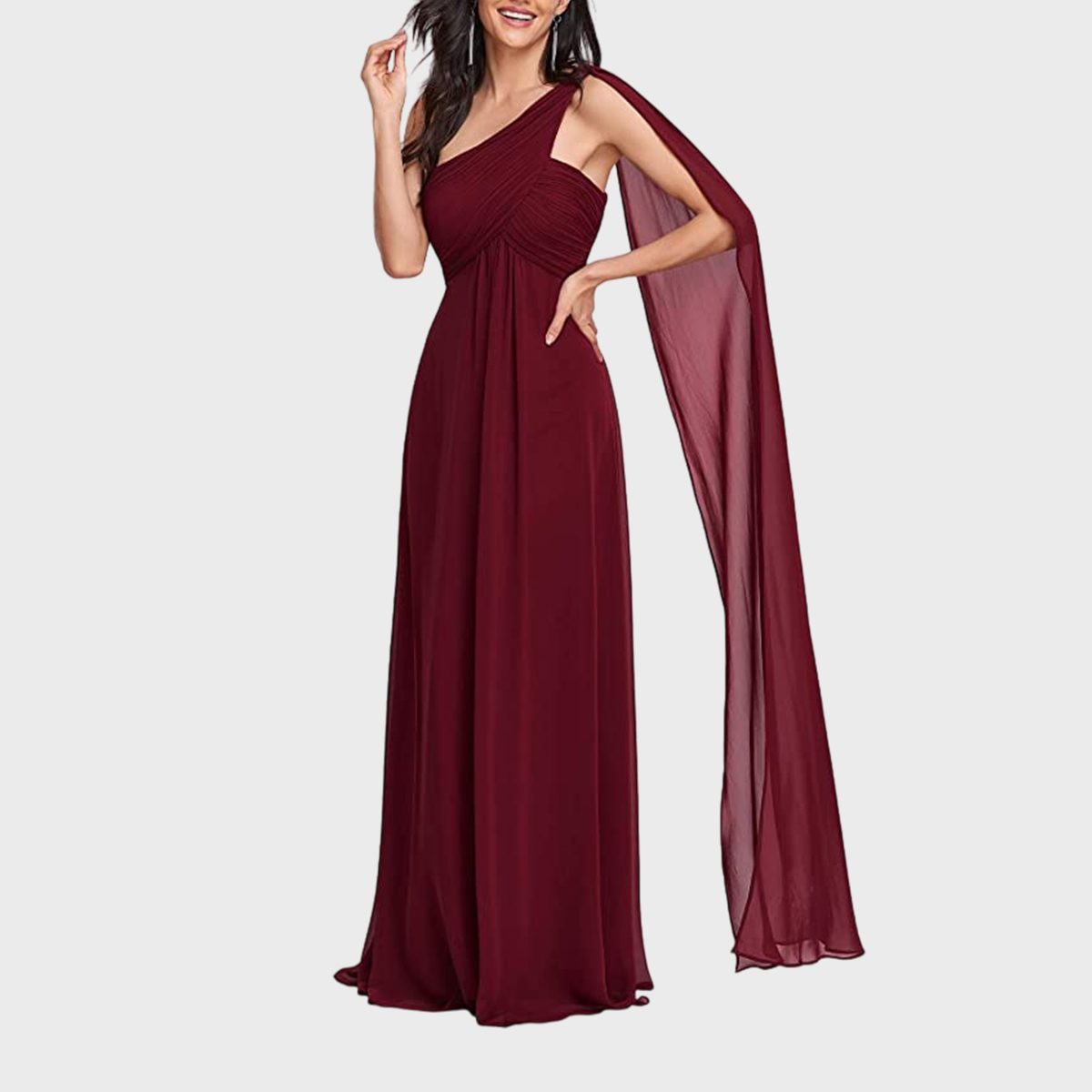 A woman wears a maroon, one-shoulder gown with a flowing cape, standing against a plain gray background.