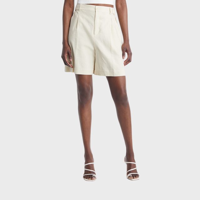 A person stands wearing beige shorts and white strappy sandals against a plain white background.