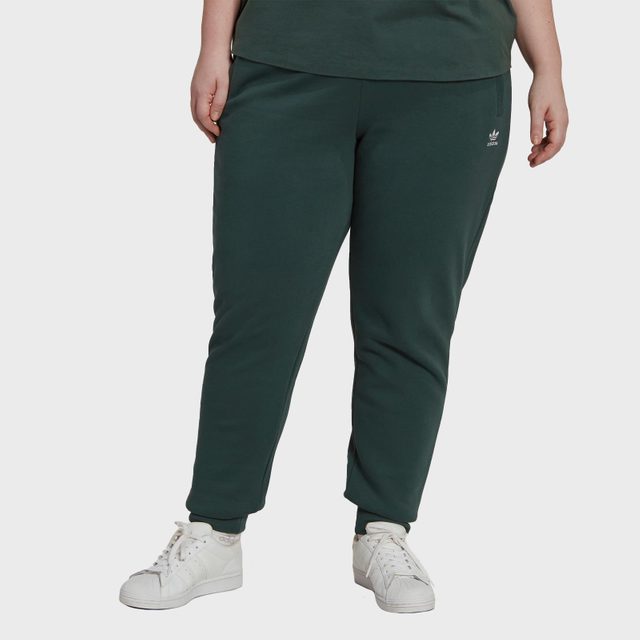 Person wearing dark green sweatpants stands on a white background; Adidas logo visible on pants and wearing white sneakers.