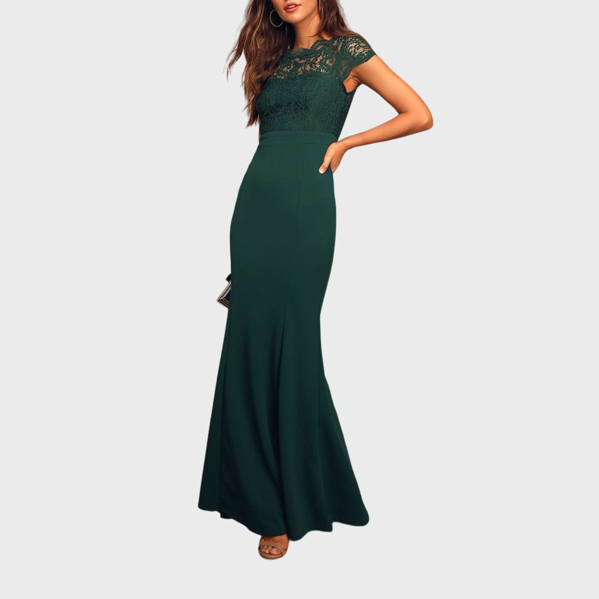 A woman stands wearing a long, elegant green dress with lace detailing, holding a clutch, against a plain background.