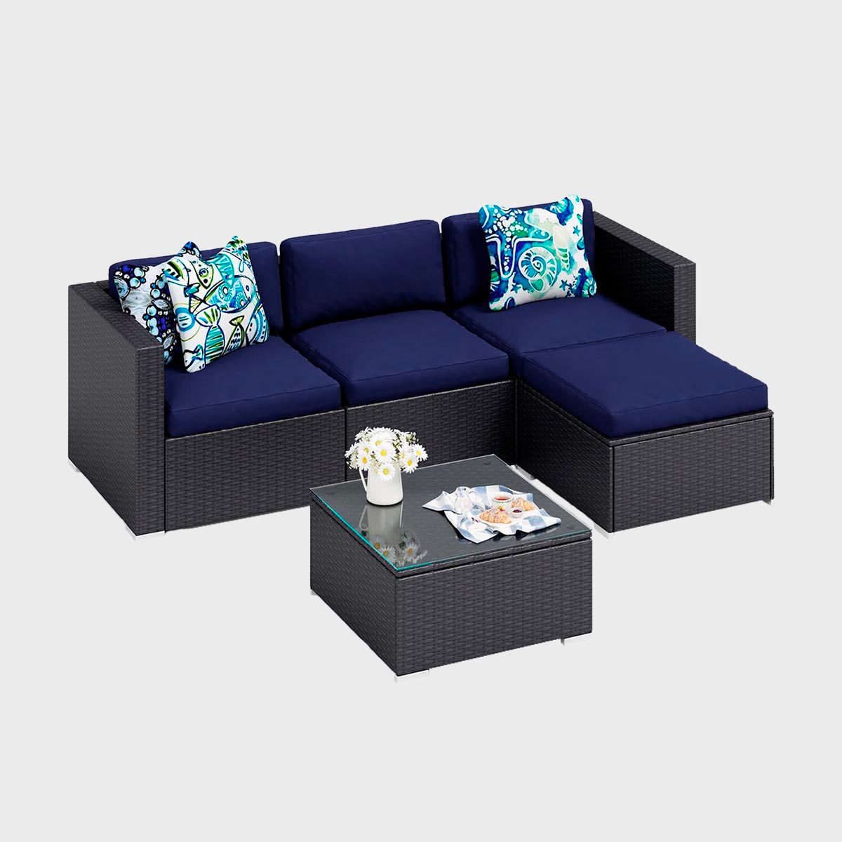 Sectional sofa sits with decorative cushions, beside a glass-topped coffee table holding a vase of daisies and a tray with snacks, on a plain background.
