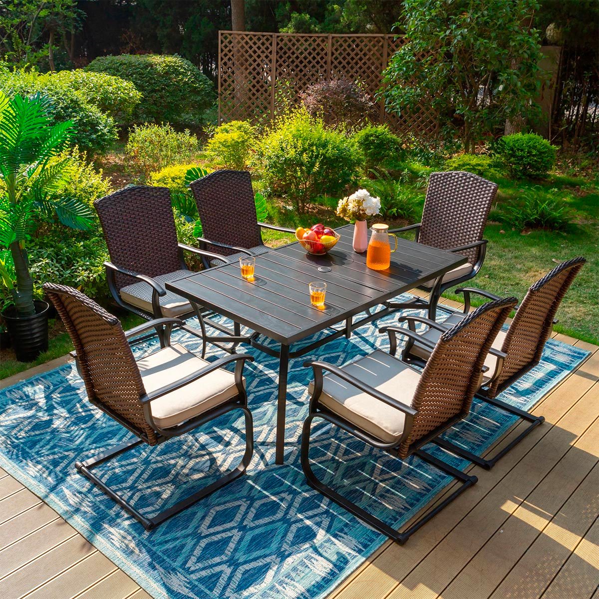 A patio set with six wicker chairs surrounds a table with drinks and flowers on a blue pattern rug, amidst garden greenery.