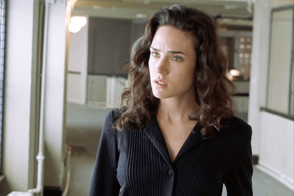 A woman with wavy hair stands in a hallway, looking intently. The background is softly lit, highlighting architectural details.