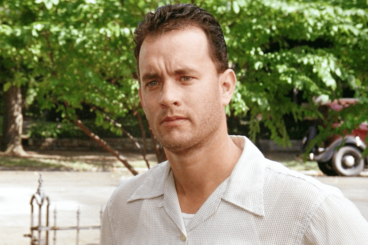 A man with short curly hair stands calmly outdoors, surrounded by green trees and a vintage car is visible in the background.