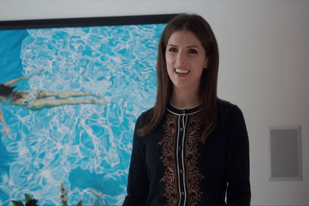 A woman smiles, standing indoors, with a framed painting of a swimmer in a blue pool in the background.