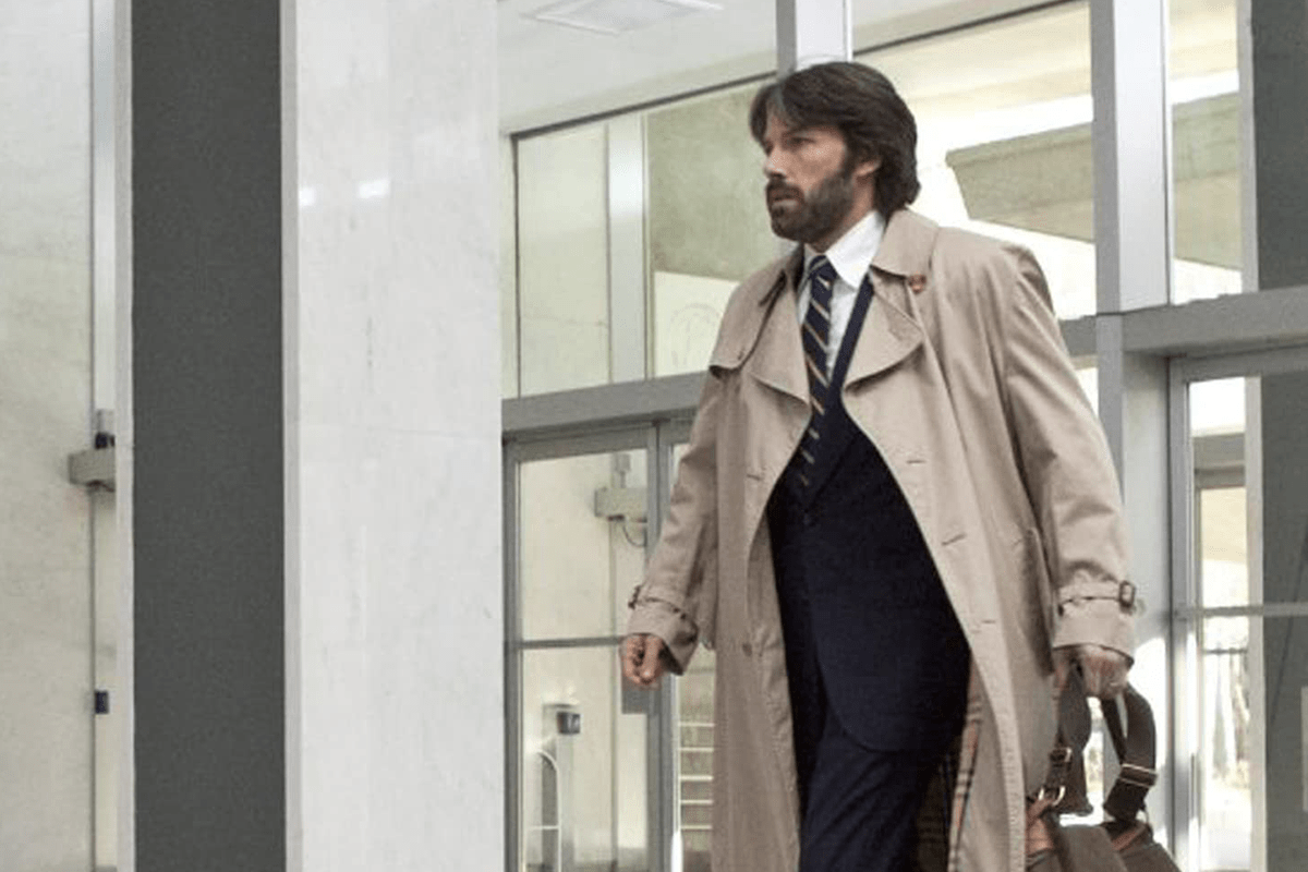 A man in a trench coat walks purposefully, carrying a bag, in a modern building with glass walls and marble surroundings.