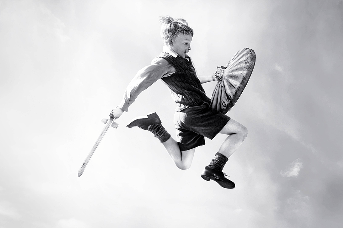 Boy jumps with wooden sword and shield, wearing shorts and sweater; background is a cloudy sky.