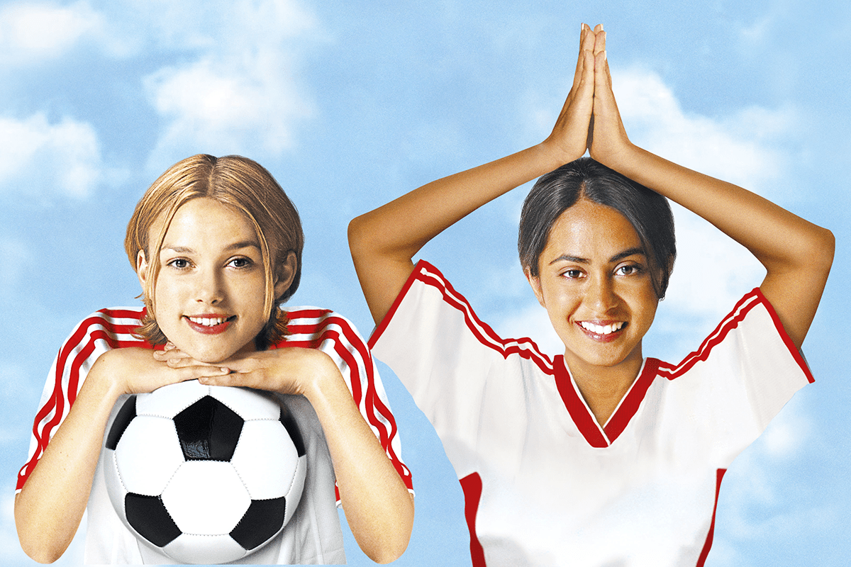 Two young women in soccer jerseys, one holding a soccer ball, the other with hands together above her head, against a clear sky.
