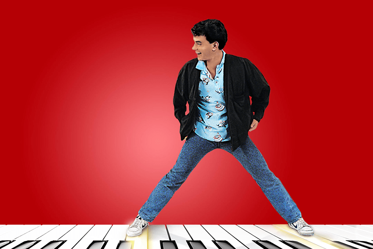 Man standing with legs wide on giant piano keys, wearing a patterned shirt and jacket, against a red background.