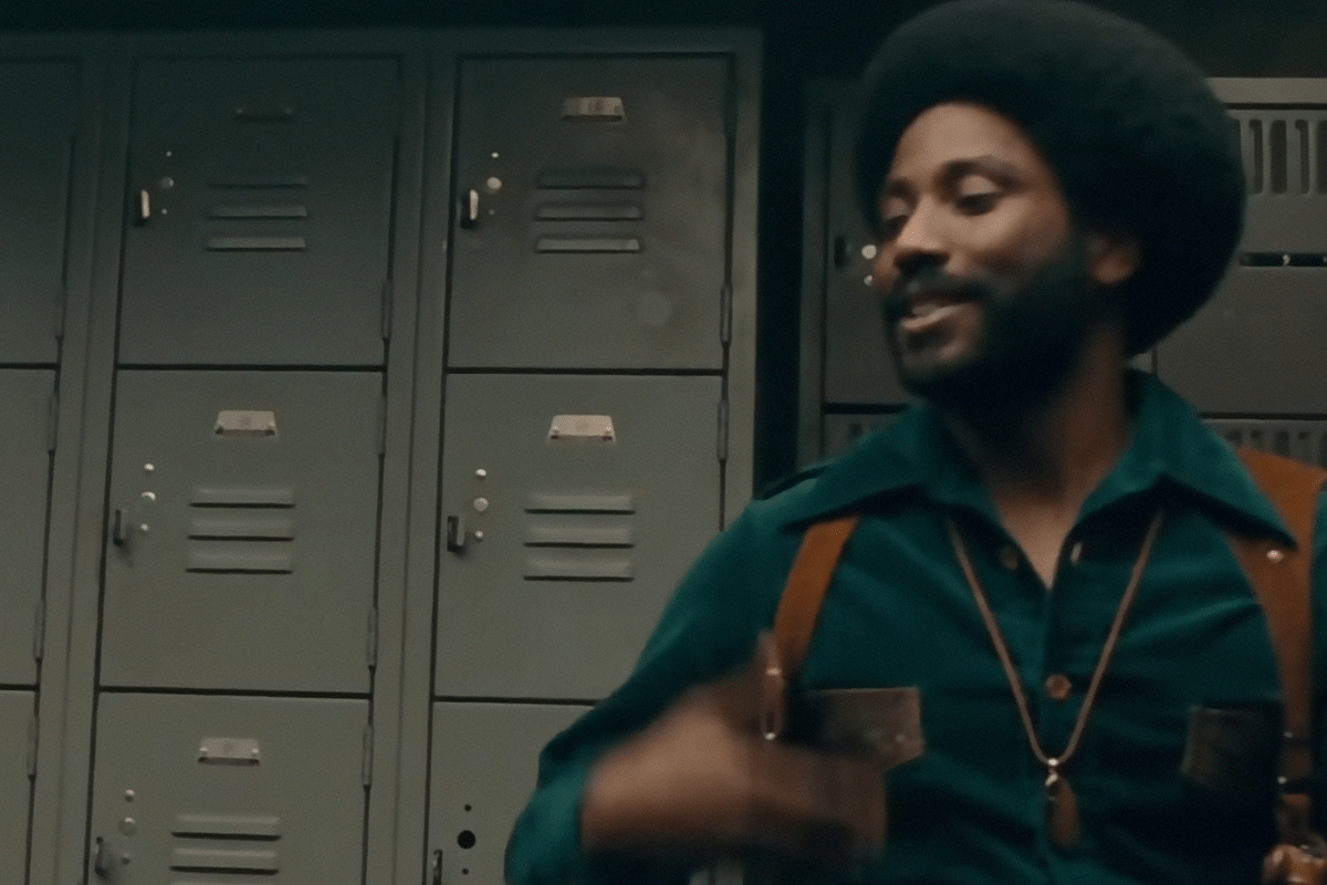 Man smiling, standing in front of a row of gray lockers, wearing a dark green shirt with suspenders.