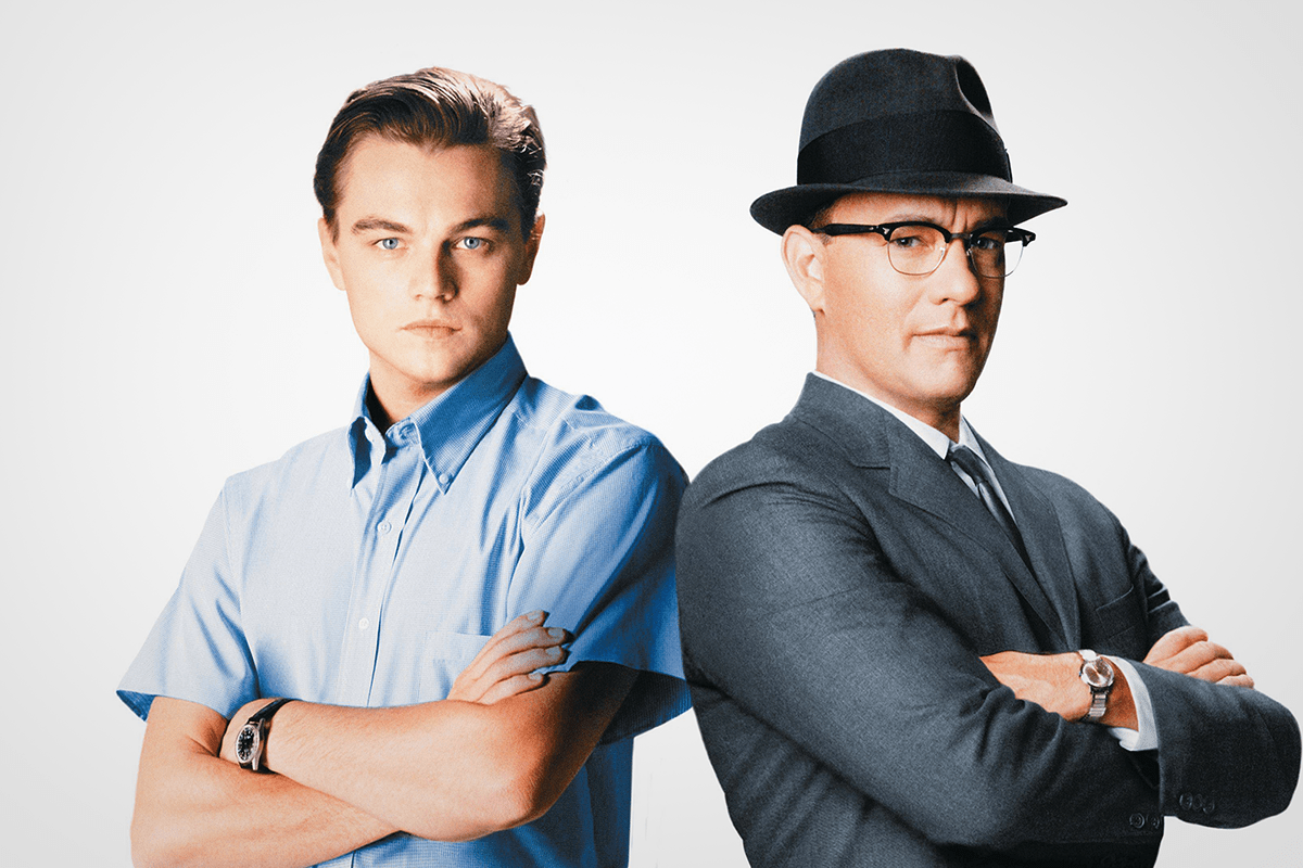 Two men stand confidently, arms crossed; one in a blue shirt, the other in a suit and hat, against a plain background.