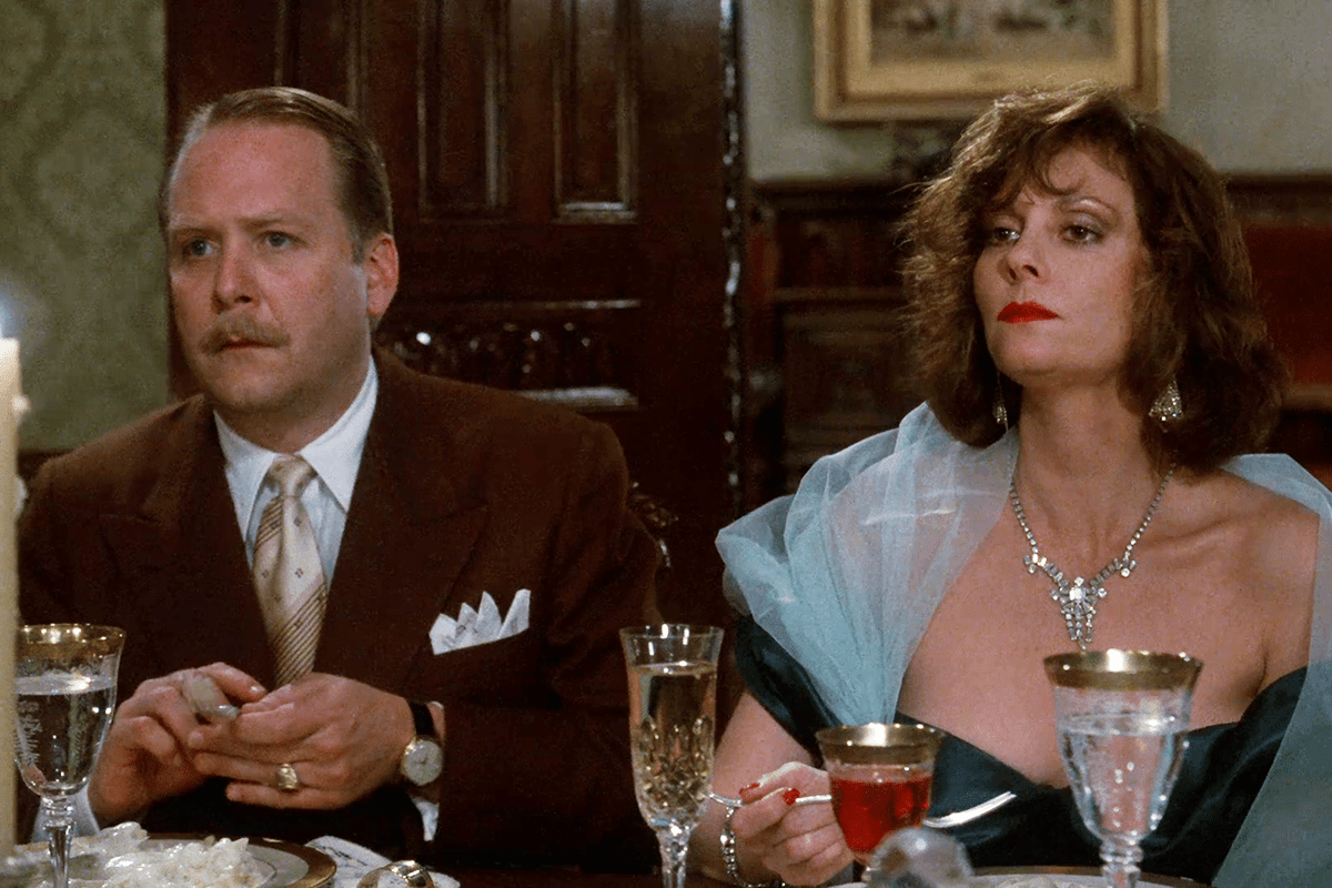 Two elegantly dressed individuals sit at a formal dining table, holding drinks, with ornate glassware and wood paneling in the background.