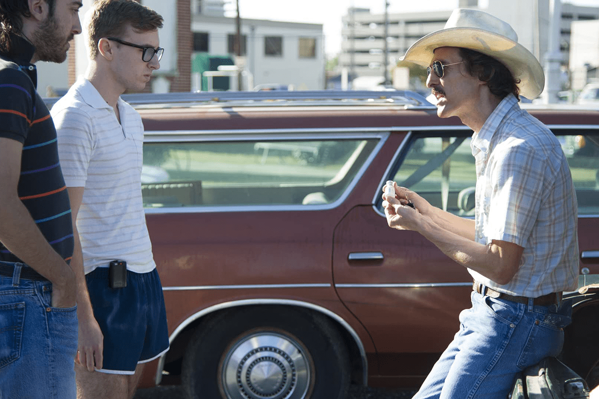 Man holding items, talking to two others, leaning against a car in a parking lot.