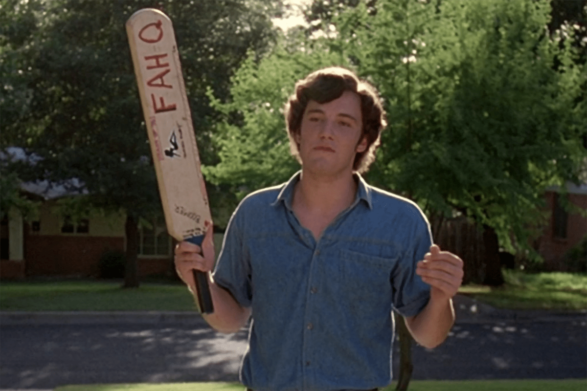 A person holds a cricket bat with "FAHQ" in a leafy suburban area, wearing a blue shirt, looking relaxed.