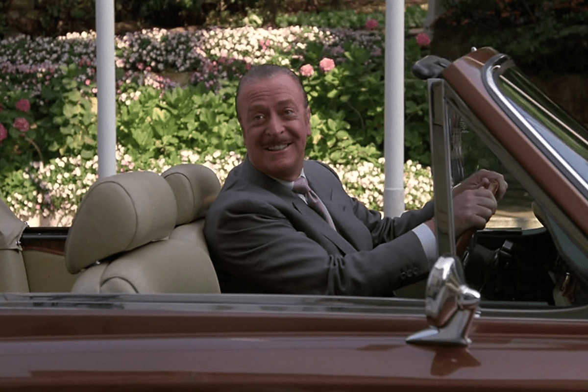 Man drives convertible, smiling, in a garden with abundant flowers and greenery.