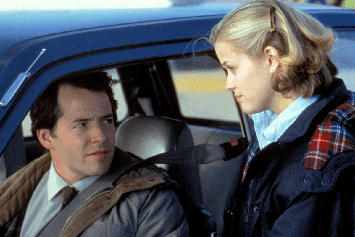 Man seated in car, looking at standing woman beside car, both wearing jackets, daylight setting.