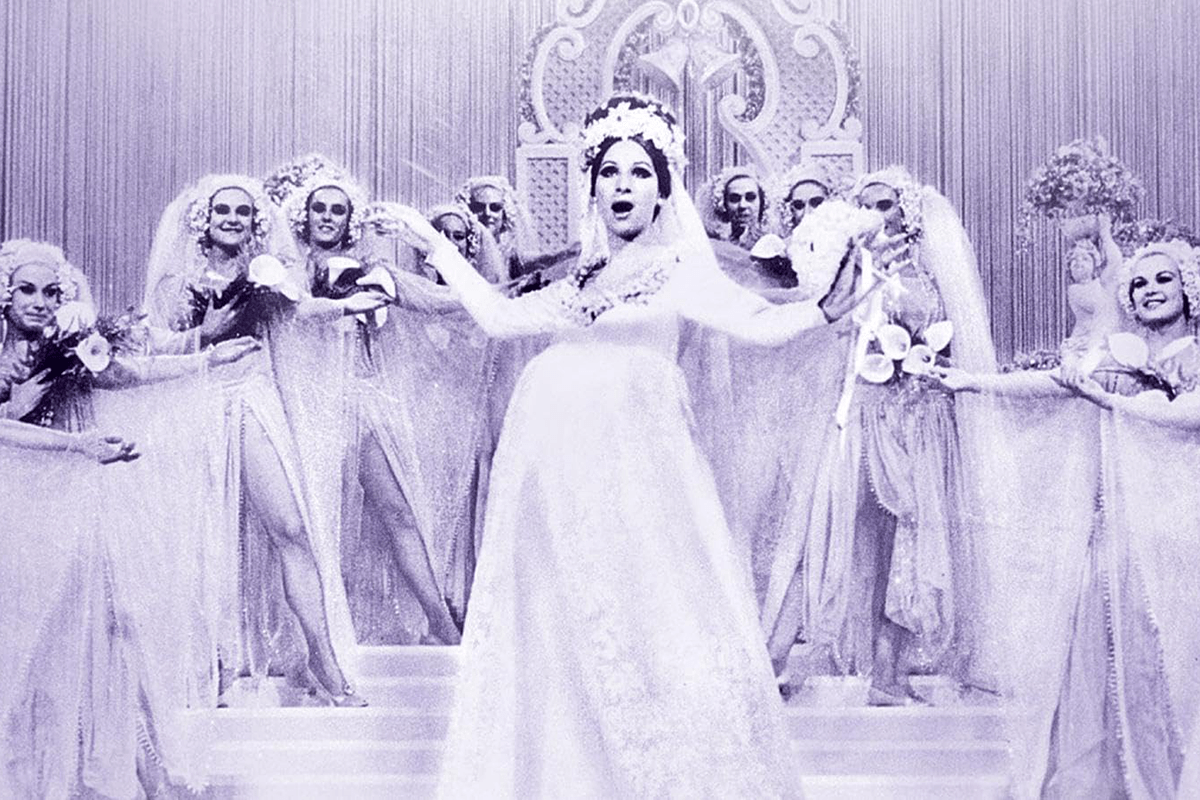 Bridal-clad woman sings onstage, surrounded by dancers with flowers, in an ornate theater setting.