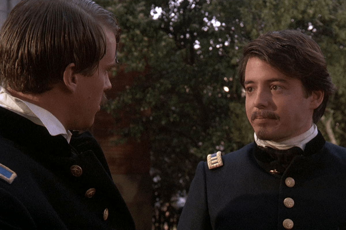 Two men in historical military uniforms stand outdoors, engaging in conversation, with trees visible in the background.