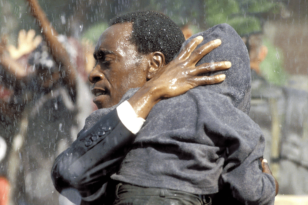 Man embraces child, shielding him amidst falling water, surrounded by blurred crowd with raised hands.