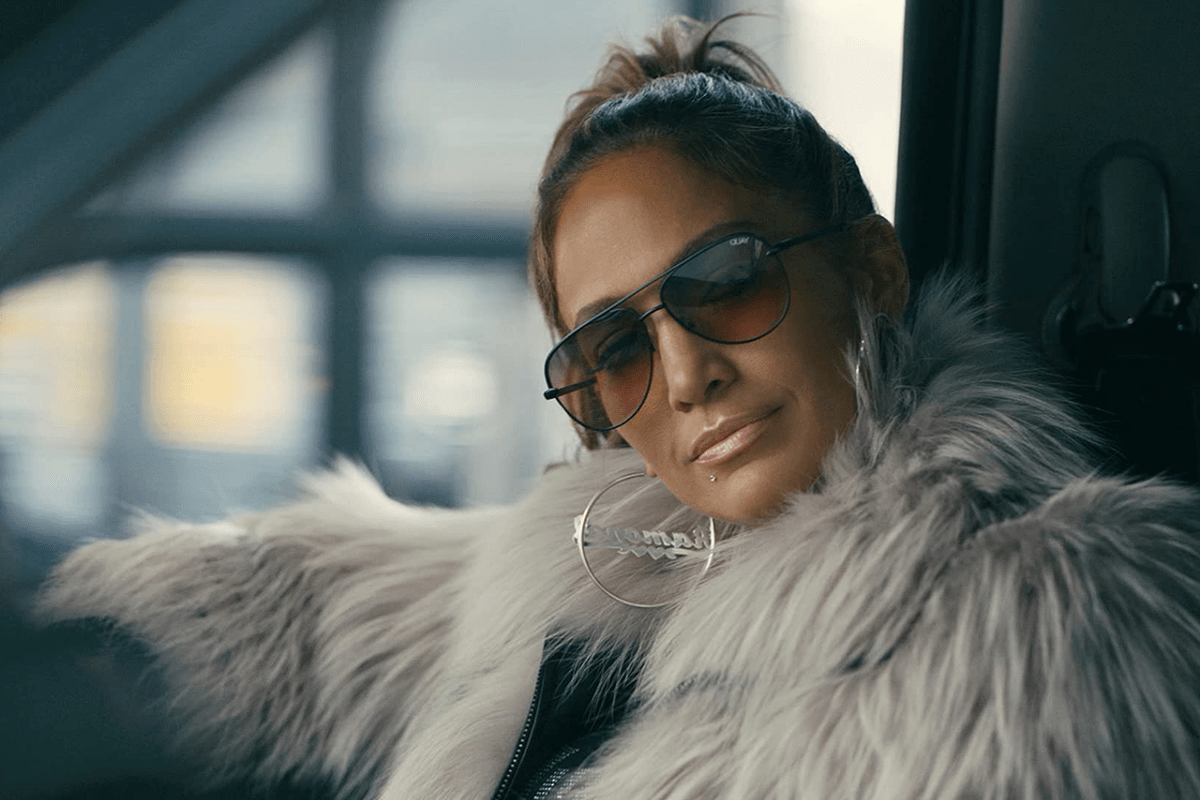 Woman wearing sunglasses and a fur coat, sitting relaxed in a car, with blurred cityscape visible through the window.