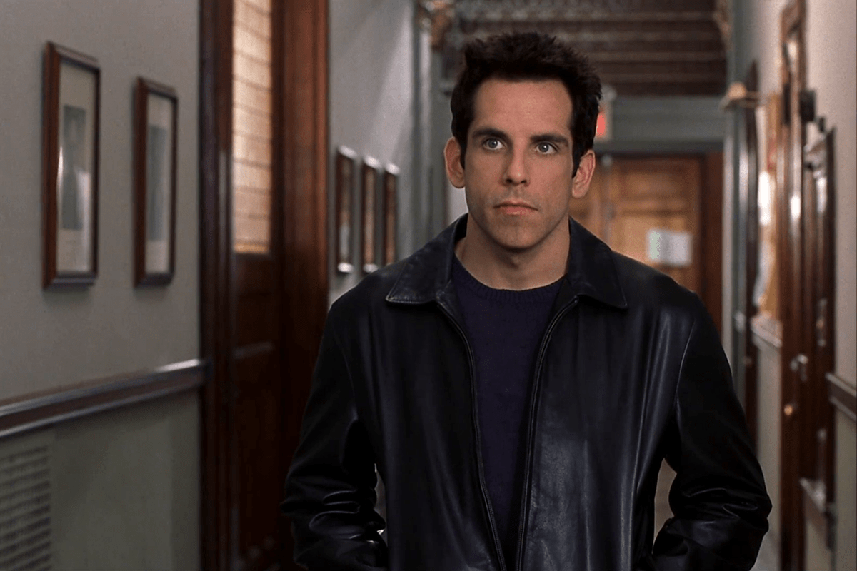 Man standing still in a hallway, wearing a dark leather jacket; framed pictures line the walls.
