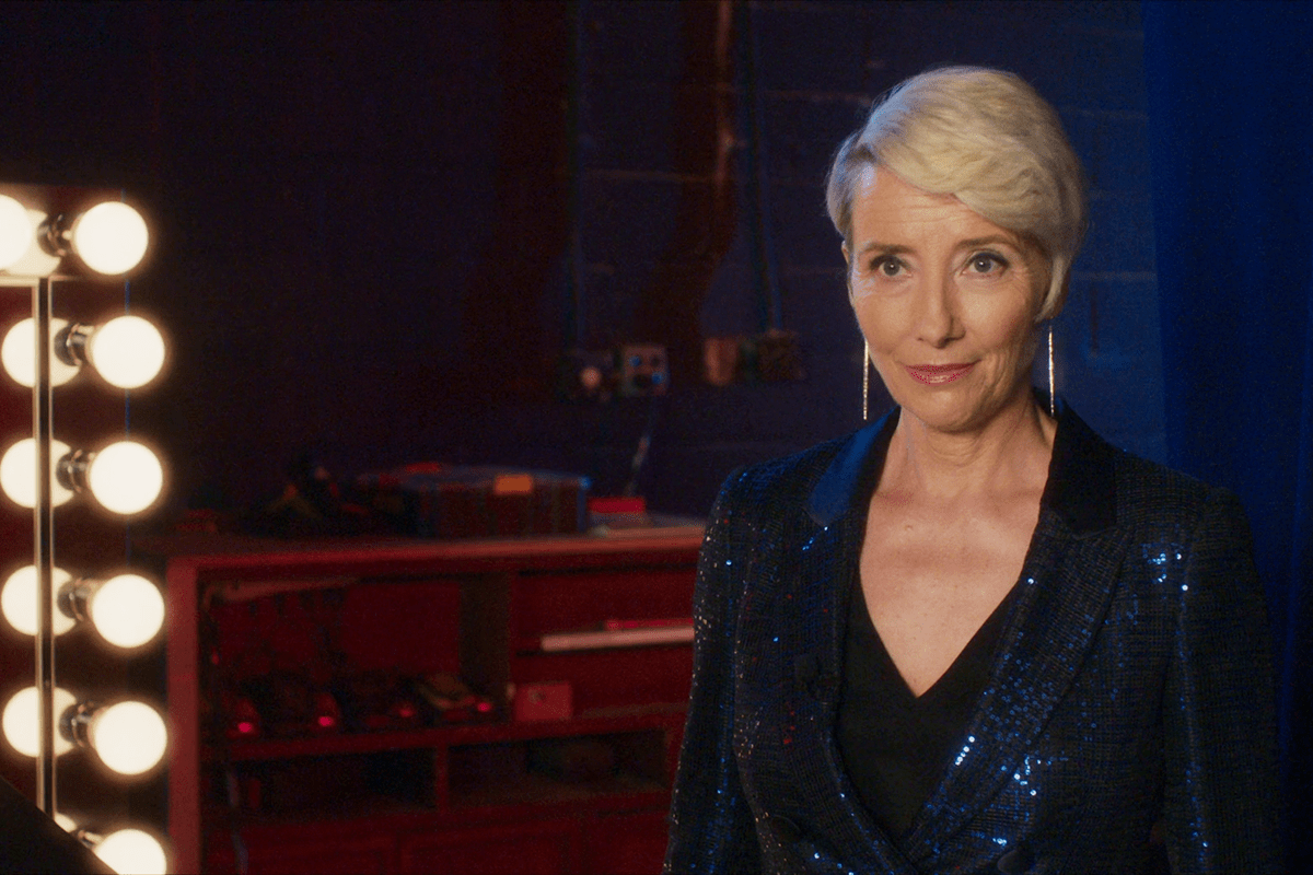 Person in sparkly suit standing confidently beside a lit mirror in a dimly lit backstage area.