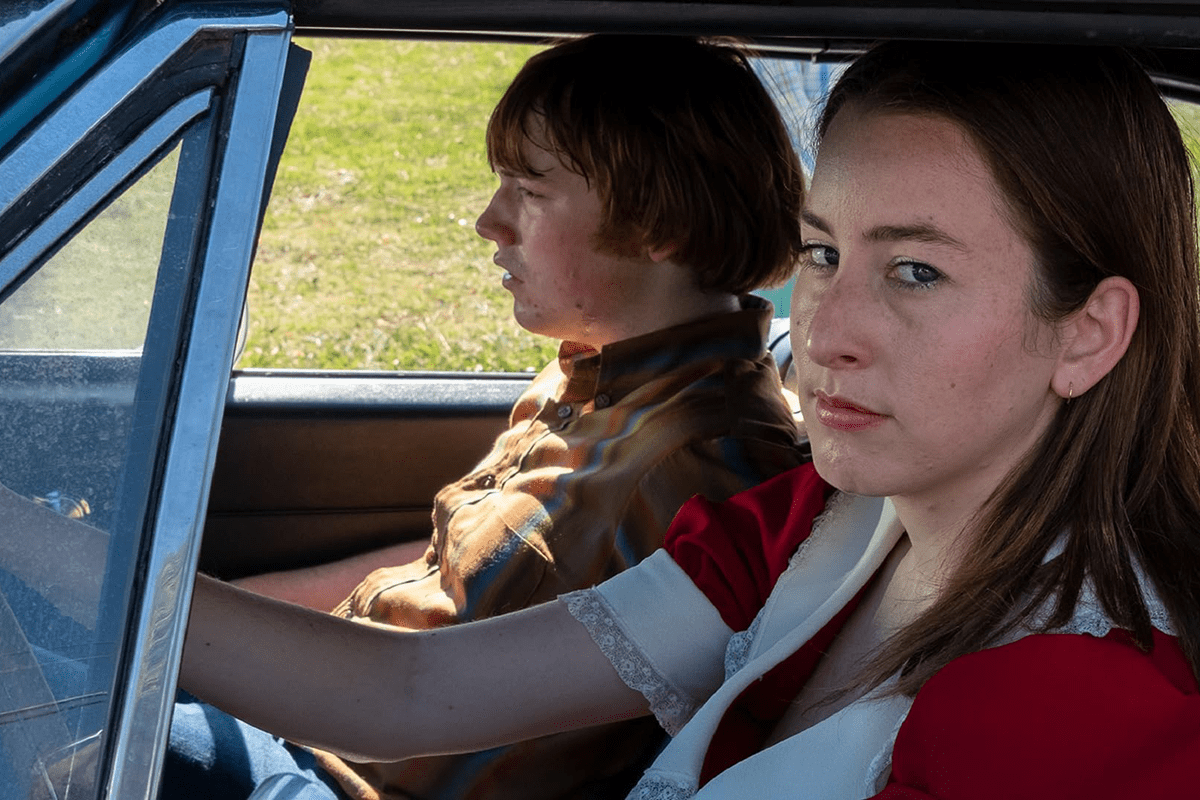 Two people sit in a car; the driver looks focused, while the passenger appears contemplative. The car is on grassy terrain.
