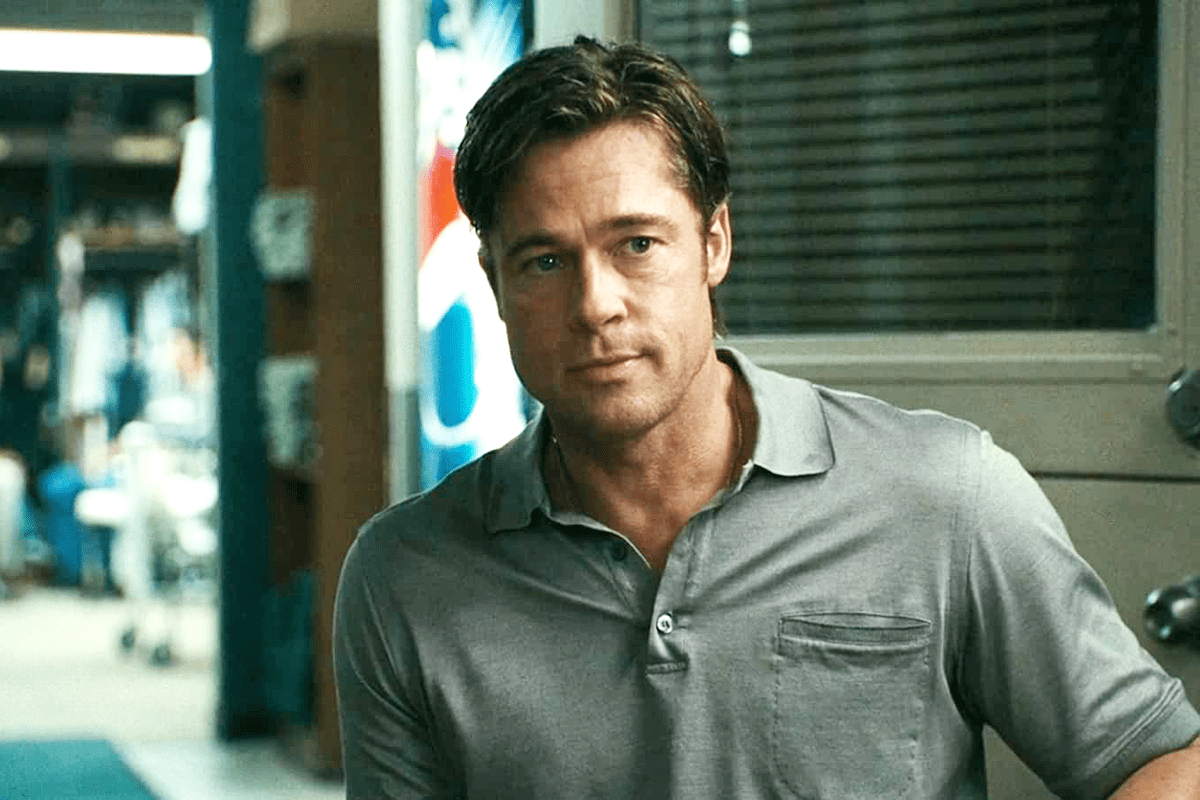 A man stands, wearing a gray polo shirt, in an office setting with blurred background and partially visible colorful posters.