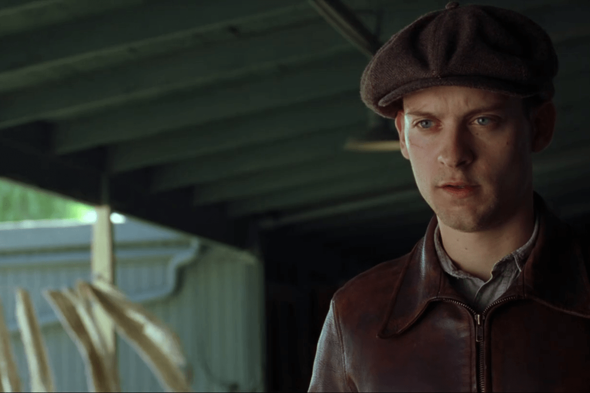 A man wearing a flat cap and leather jacket stands, appearing focused, under a wooden structure with beams and corrugated metal visible.