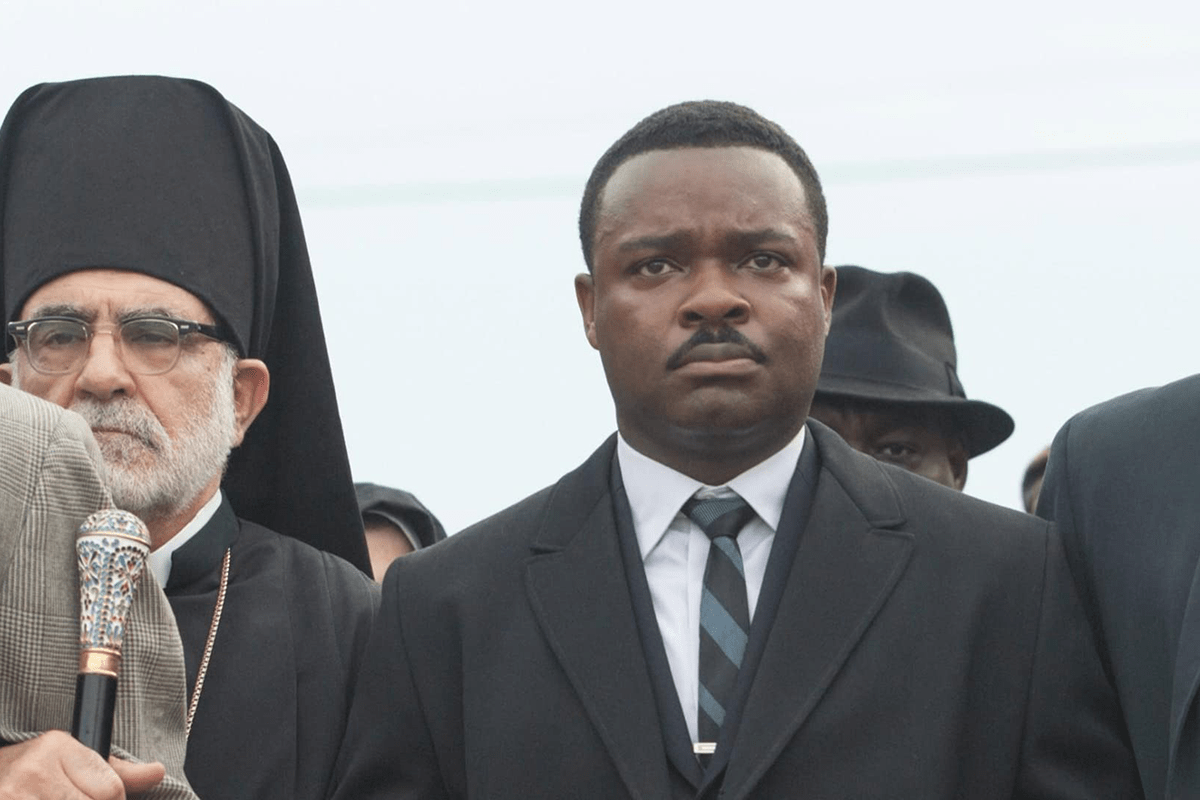 A serious man stands, wearing a suit and tie, among people dressed formally, outdoors under a cloudy sky.