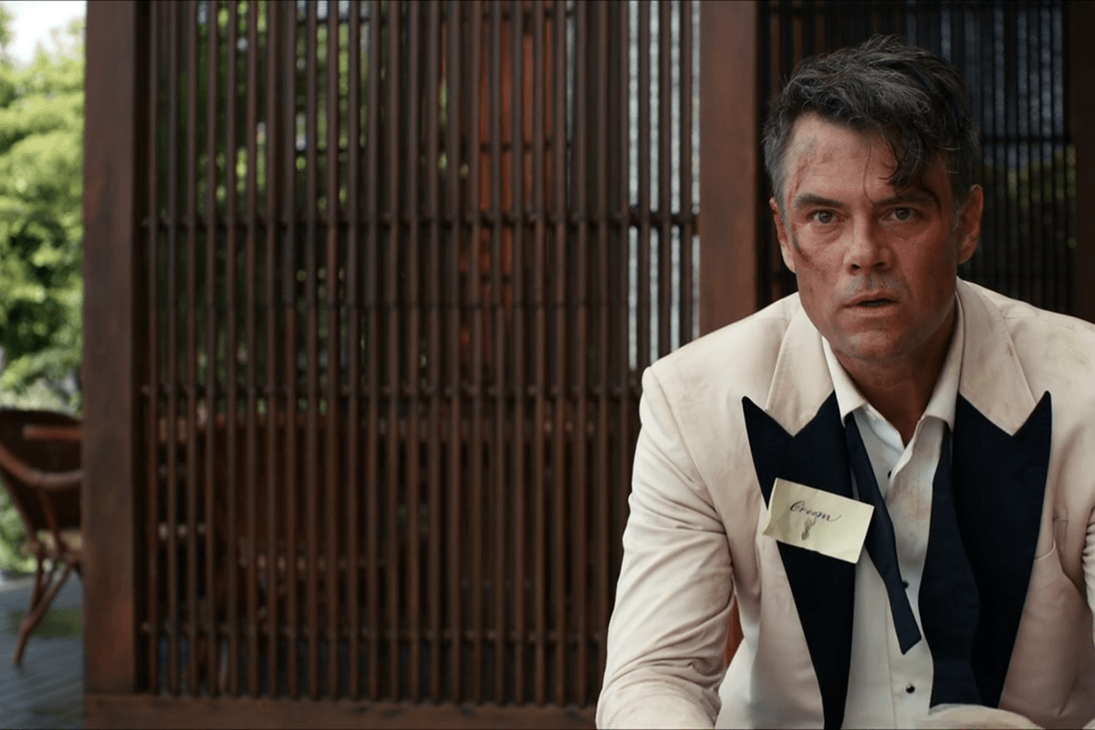 A man sits disheveled, wearing a tuxedo with a name tag reading "Groom," against a wooden backdrop and lush greenery outside.