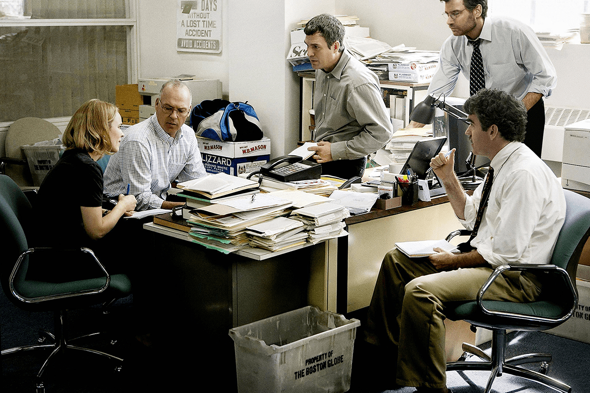 Five people discuss at a cluttered office table, surrounded by stacks of papers and boxes, with a Boston Globe recycling bin nearby.