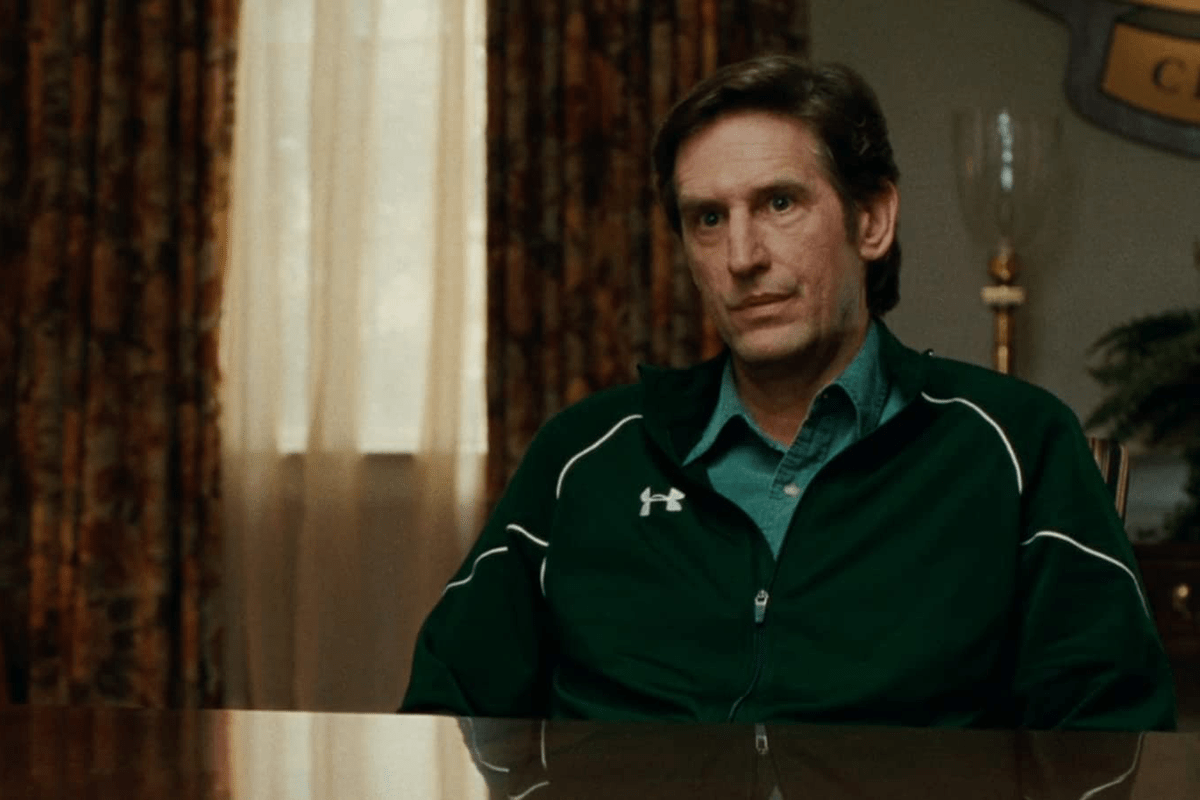 A person in a dark green jacket sits at a reflective table, with patterned curtains and soft lighting in the background.