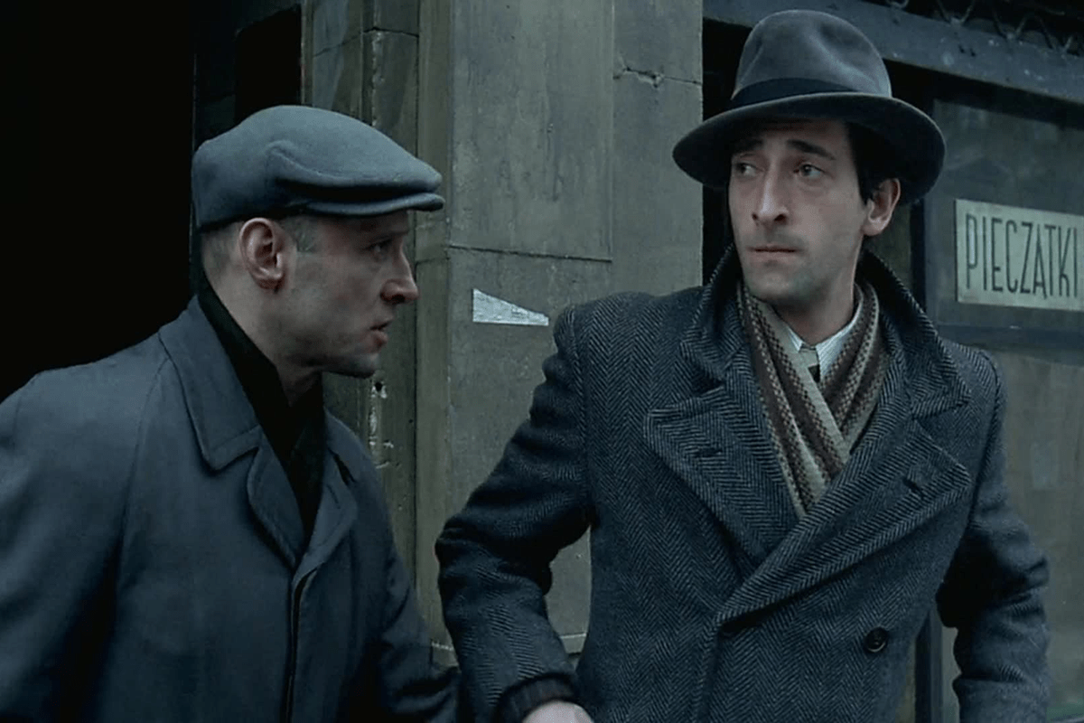 Two men stand close, wearing coats and hats, in front of a building. A sign reads "PIECZ&Auml;&bdquo;TKI" in the background.