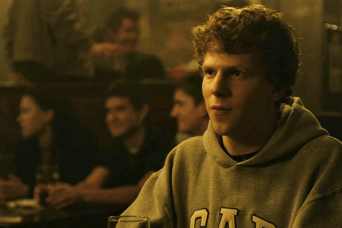 Person sits quietly, wearing a hoodie, in a dimly lit bar with blurred people conversing in the background.