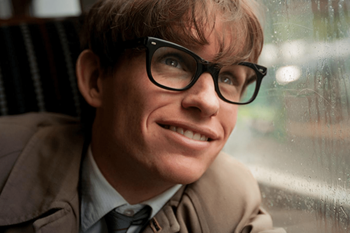 Person wearing glasses smiles, sitting near a rain-speckled window on a train, with a blurred green landscape outside.