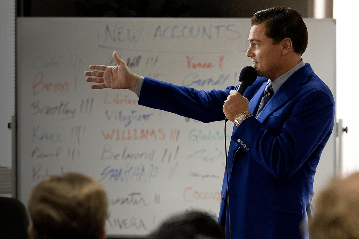 A man in a blue suit speaks into a microphone, gesturing towards a whiteboard listing new accounts, in a professional setting.