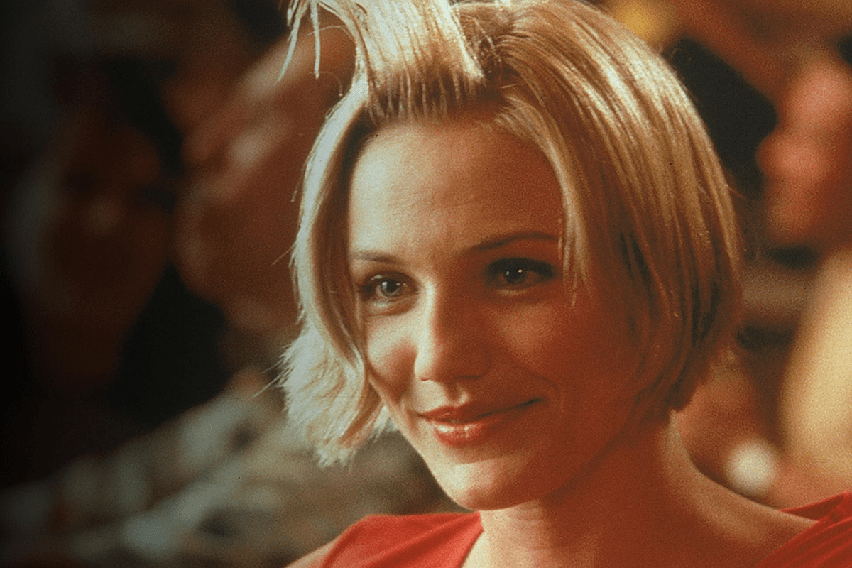 Woman smiling with hair styled upward, wearing a red top, in a dimly lit room with blurred people in the background.