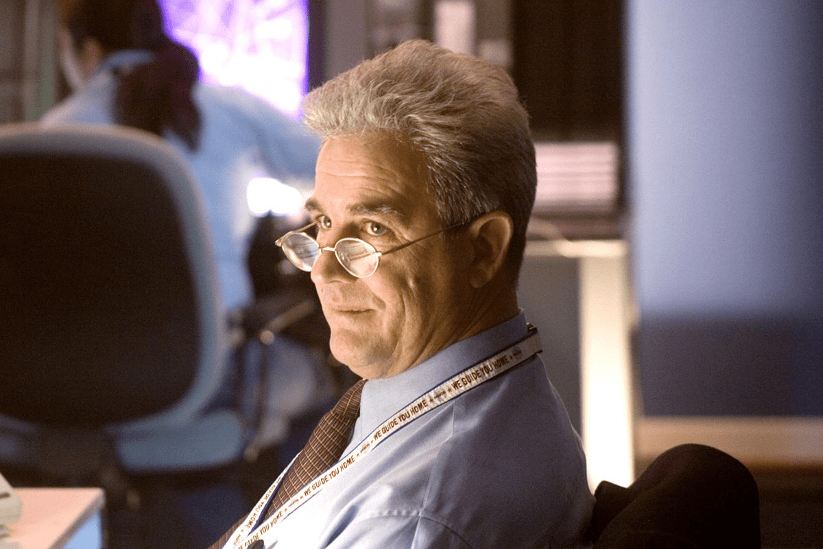 Man sitting, wearing glasses, looking sideways in an office. A lanyard reads "WE GUIDE YOU HOME." Another person blurred in background.