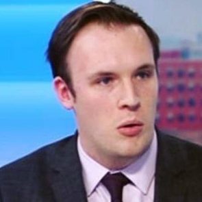 A person speaks, wearing a suit, against a blurred background featuring blue and red hues.