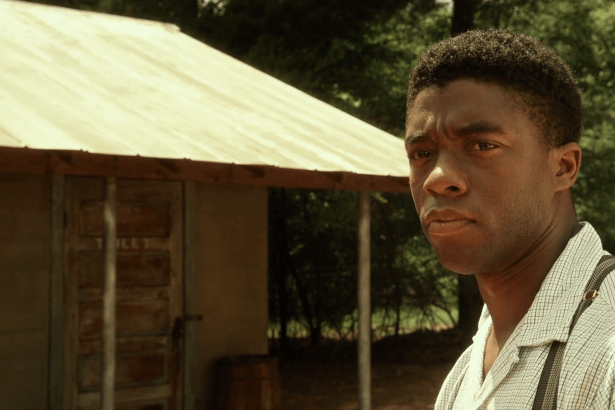 A person stands outside a weathered building with a slanted roof, labeled "TOILET," set against a background of trees.